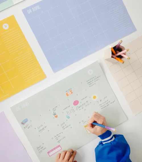 Persona escribiendo en calendario ilustrado de The Full Planner sobre mesa con planificadores de colores