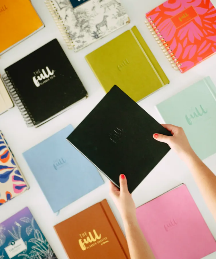Manos sosteniendo planner negro de The Full Planner sobre mesa con cuadernos de colores